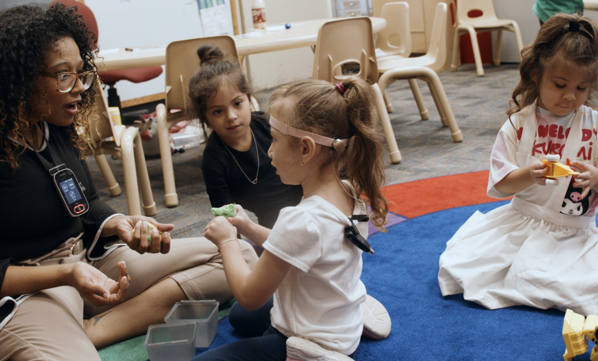 Lauren Fleming sits on a carpet with three children. She is talking with one child about their play as another child looks on. A third child is playing with trucks.