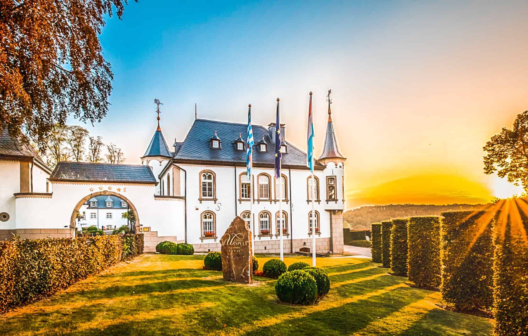 Ch&acirc;teau d'Urspelt, a grand white historic castle with turrets and flags under a sunset sky