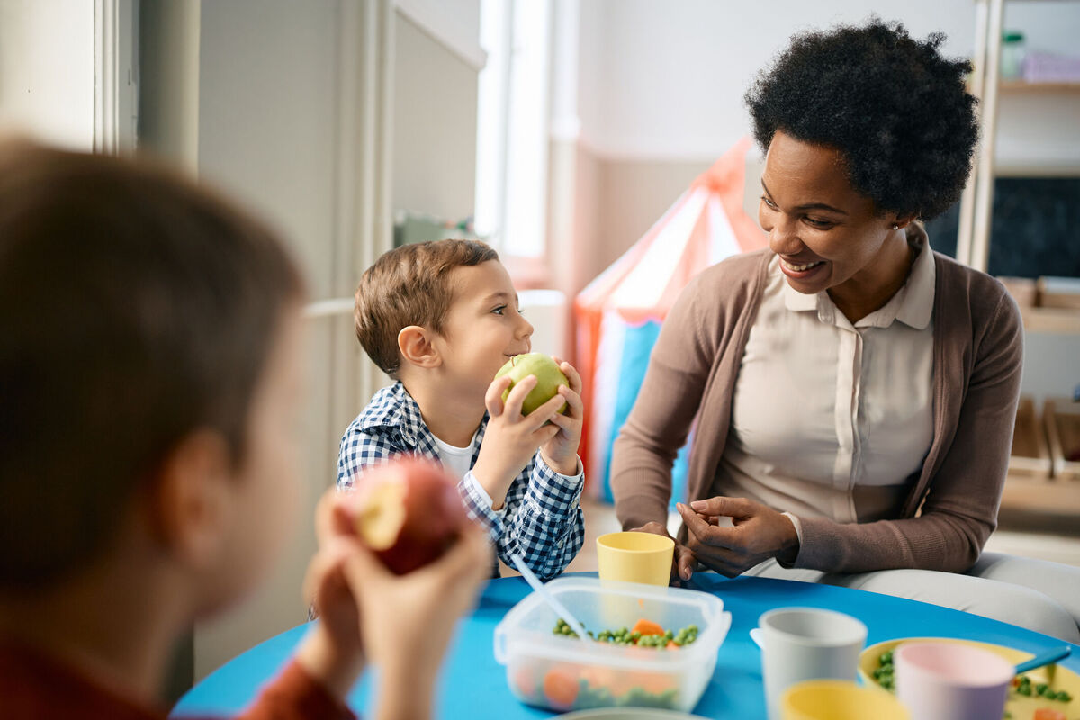A preschool teacher talks with children at lunchtime about the apples and other healthy food they're eating.