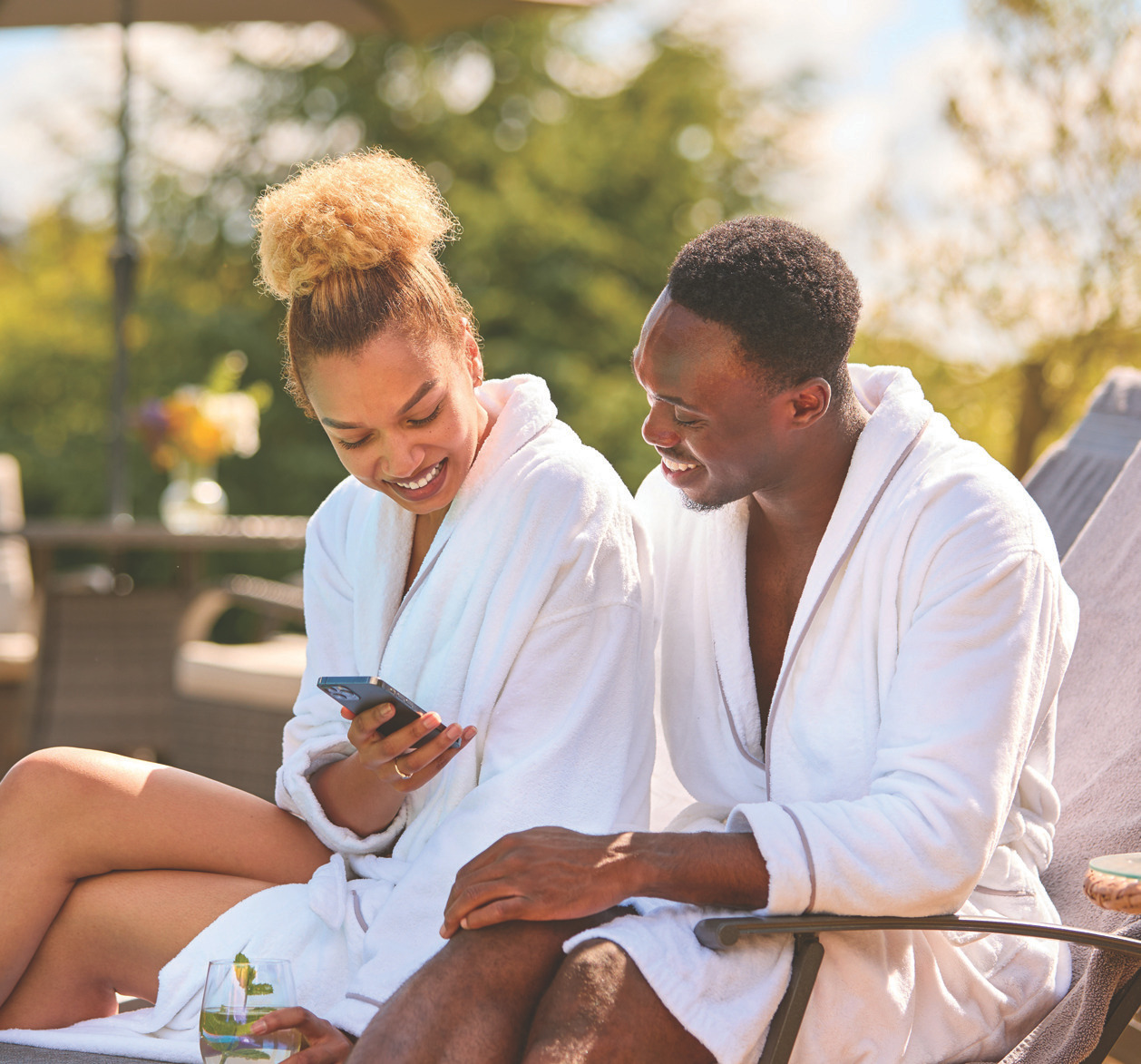 Two people in white bathrobes outdoors looking at a smartphone in a spa-like setting