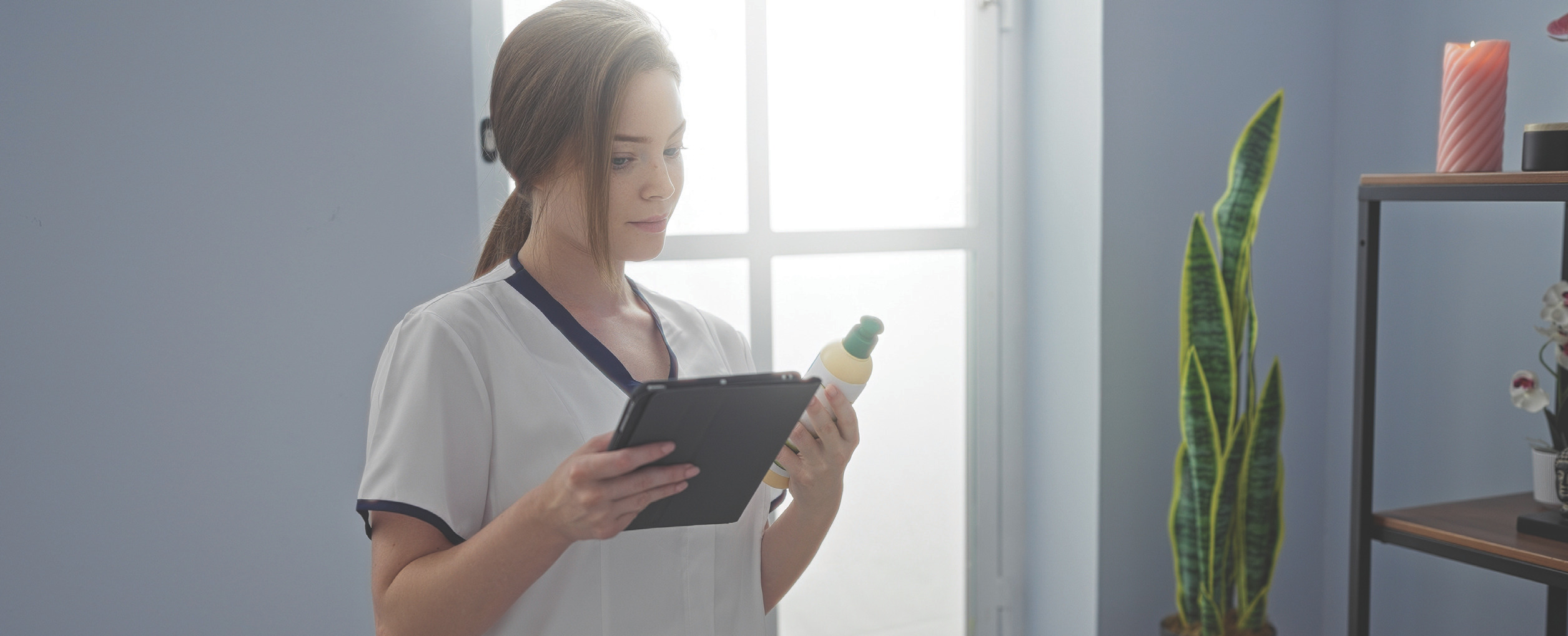 Spa therapist standing by a bright window holding a tablet and a product bottle.