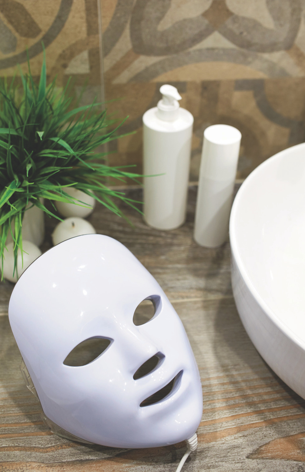 LED facial mask and skincare bottles on a wooden surface beside a sink and plant.