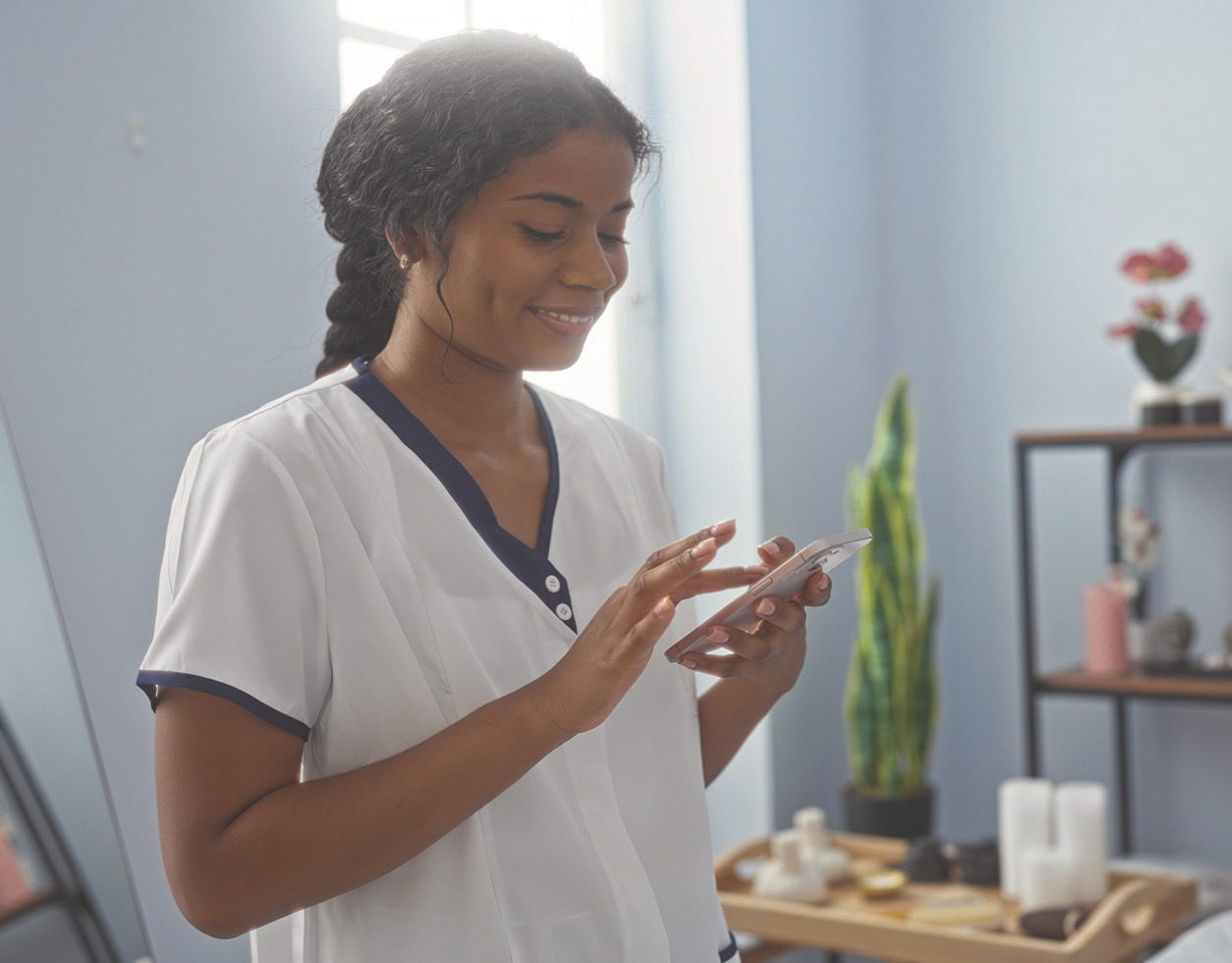 Smiling spa professional in uniform checking a smartphone in a treatment room.