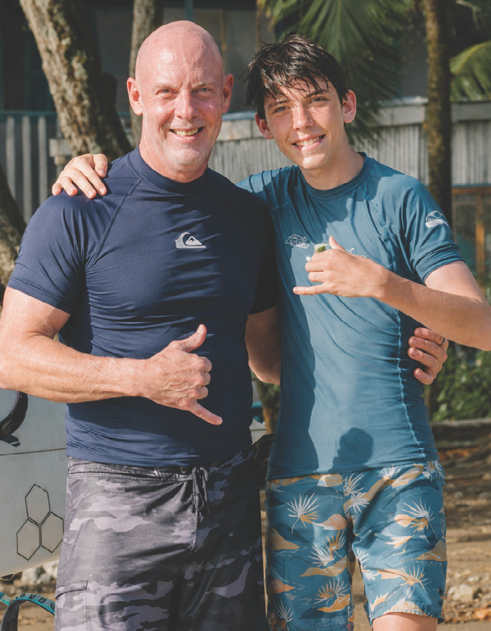 Photograph of Kenneth and his son wearing surfing rash guards and board shorts, standing outdoors and making the shaka hand sign with cheerful expressions.