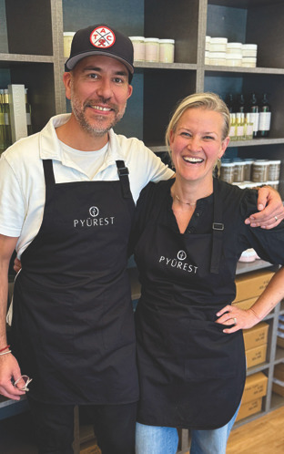 Two Py&uuml;rest Mineral + Mud staff members smiling together in aprons inside a retail/spa setting.