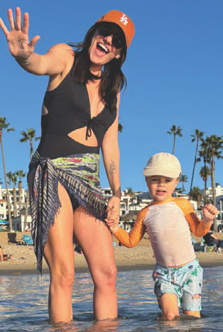 Woman with a small child standing in shallow ocean water at the beach, smiling and waving.