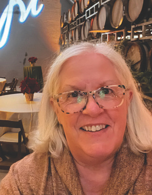 Portrait of an older woman with glasses smiling in a dining or tasting-room setting.