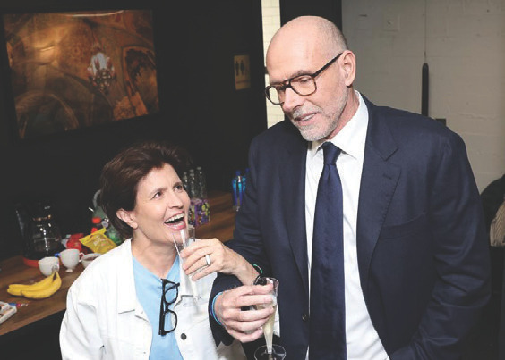 Two older adults at an event: a woman laughing while holding a drink and a balding man in a suit beside her