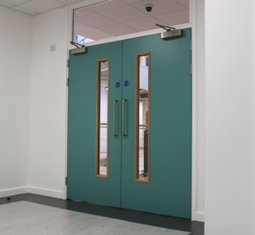 Teal double doors with narrow vertical vision panels in a corridor, showing installed electromagnetic door closers above each door.