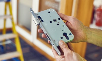 Hands holding a mortice lock case, showing the front face and fixing holes.