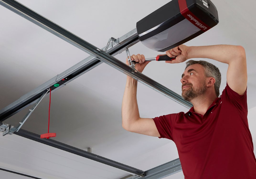 Technician installing a LiftMaster garage door opener on the ceiling using a screwdriver; red emergency release cord visible.