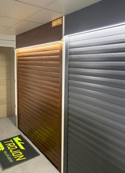 Showroom display of two roller doors with integrated LED strip above left wood‑grain door