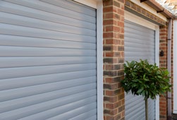 A pair of light grey roller garage doors with brick surrounds and a small plant beside them.