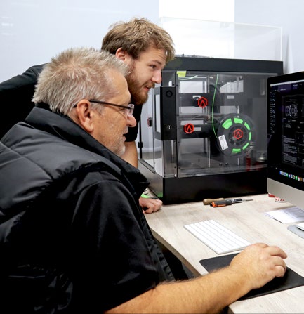 Two technical team members working at a desk beside a clear acrylic enclosure and a computer screen