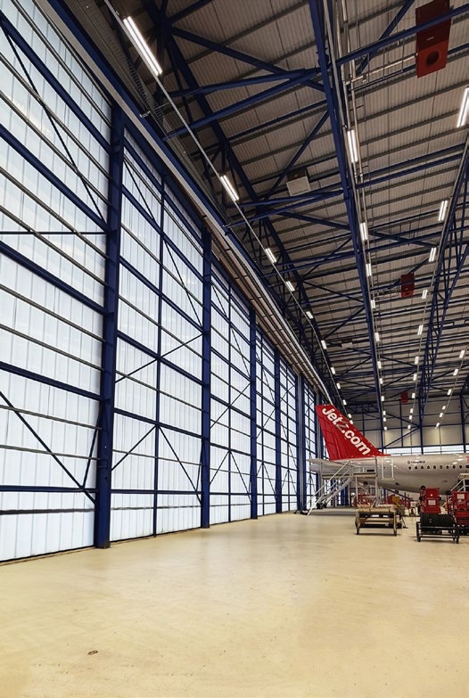 Interior view of a large aircraft hangar with high polycarbonate panel walls letting in natural light; a Jet2 aircraft is partially visible