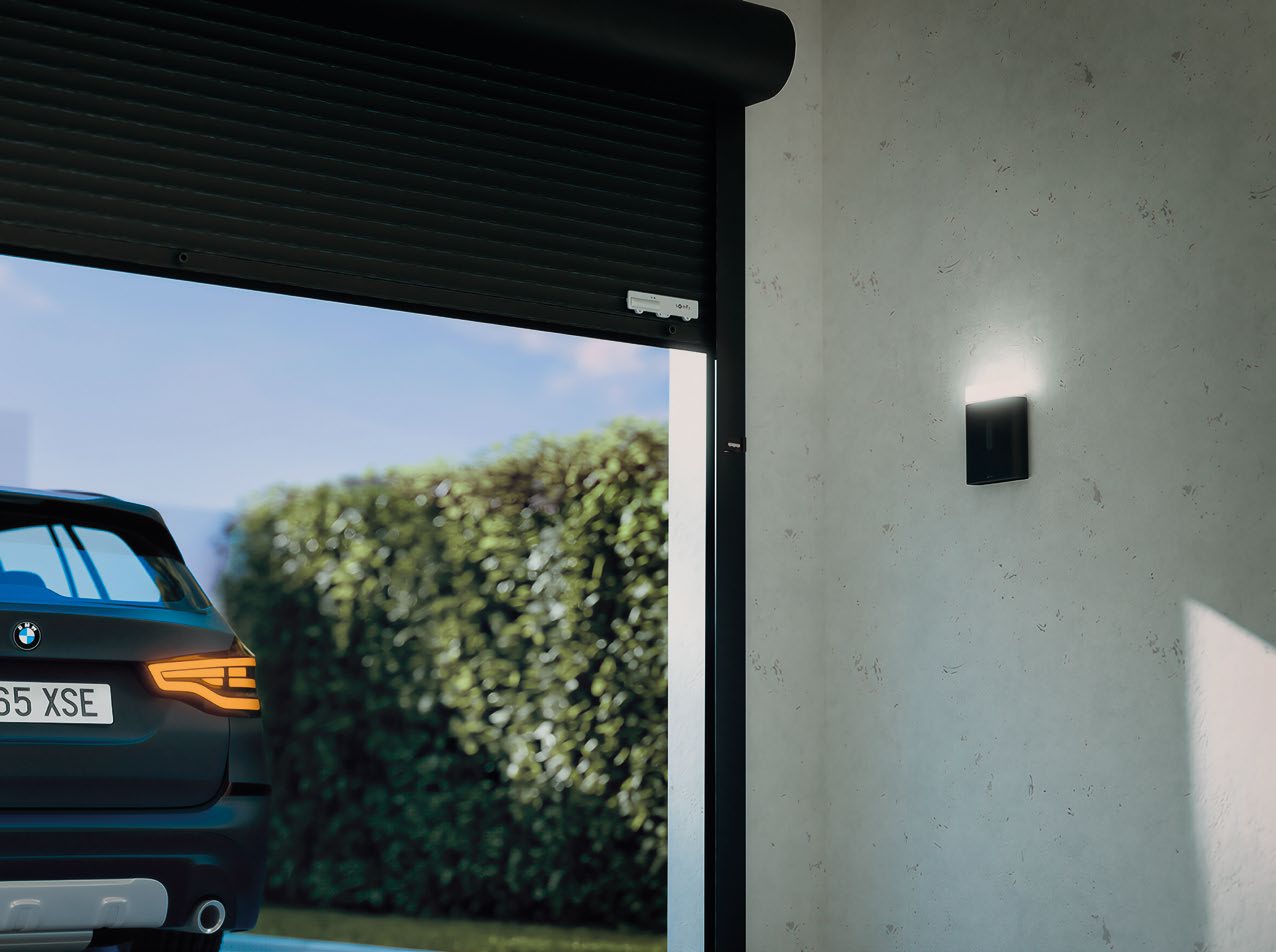 Garage interior with roller door partially open, showing a car outside and illuminated wall light beside the door.
