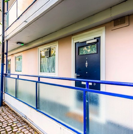 External apartment walkway showing frosted glass panels, blue metal balcony railing and a dark blue front door