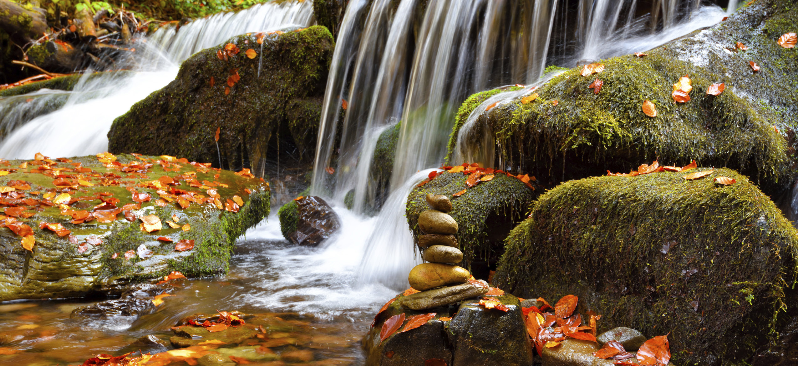 A small waterfall tumbling over moss-covered rocks with autumn leaves and a stacked cairn of stones at the foreground