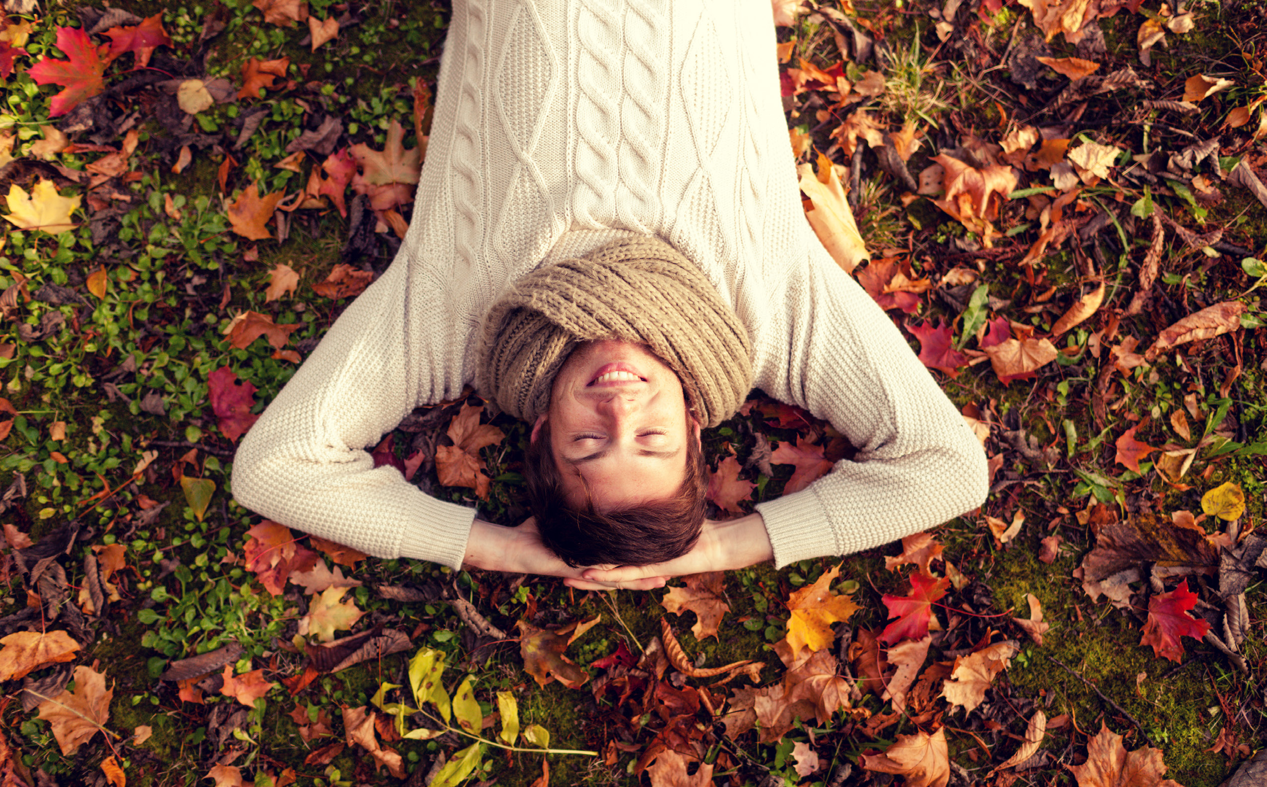Person lying on autumn leaves wearing a cream sweater and scarf, viewed from above