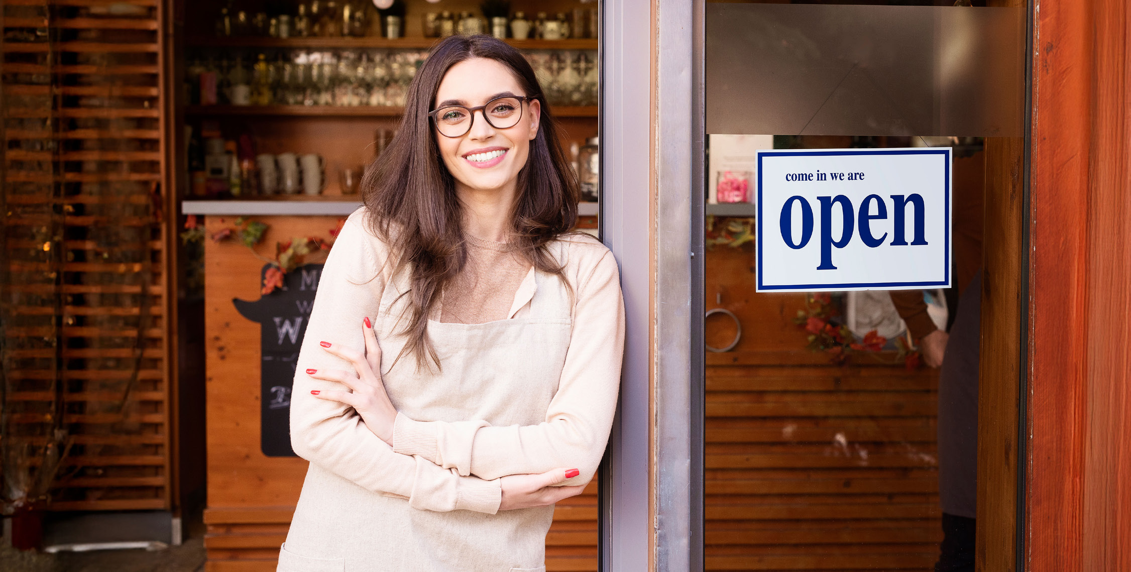 A smiling franchise owner in a white apron standing in front of an 'Open' sign at her local business storefront.